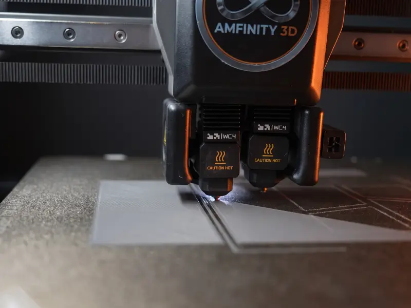 Doppeldüsen-Druckkopf mit AMFINITY-3D-Logo legt die erste Schicht eines Bauteils auf der Druckplatte ab